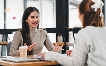 Two women having friendly conversation at table in modern office space, with one holding coffee cup and other gesturing expressively, creating collaborative and engaging moment